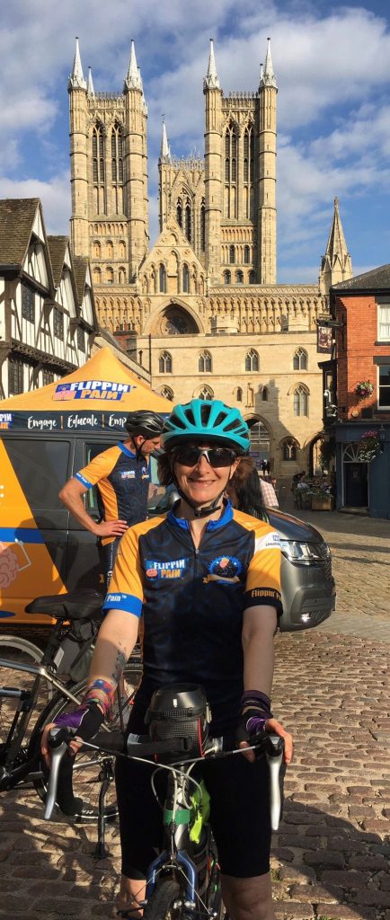 Niki in front of Lincoln Cathedral in Flippin Pain branded cycling gear and her bike smiling very happily